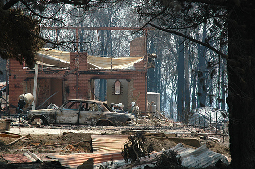 Wreckage of a house and a burned out car sit among trees.