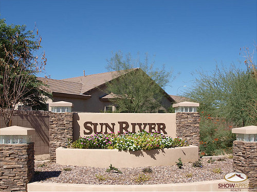A large tan home and several small palo verde trees peek out above a tan adobe and stone wall with brown letters saying 'Sun River'