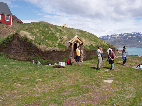 Several male tourists stand around outside a house made of sod. A red barn and snow-capped mountains can be seen in the background.