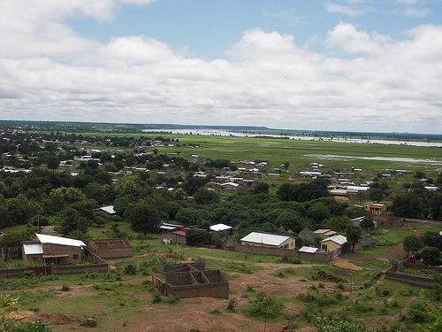 An aerial view of a village made up of small, detached houses. The ground in the background is covered in pasture leading up to a river, while the foreground is bare dirt. Some trees grow in between the houses