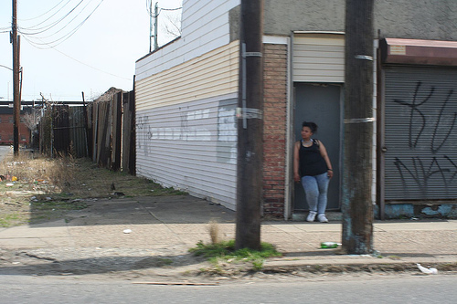 A black woman stands in the doorway of a shuttered store, whose walls are covered in graffiti. Next door is an empty lot