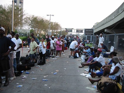 Two lines of people, most of them African American and wearing casual summer clothes, stand and sit on a stretch of pavement next to a highway overpass