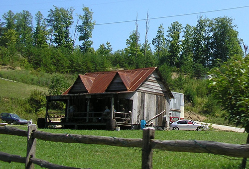 A gray wooden house sits in a large green lawn, with forest behind it. Several people sit on the porch, with a car and truck in the driveway.