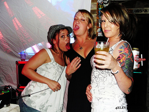 Three intoxicated white women pose in a dance club. One holds up a glass of beer, while the other two stick out their tongues and lean against each other in a sexually suggestive way