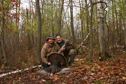 Two white men -- one much older with a white beard, the other younger with a brown beard -- kneel in the forest, posing with a rifle and a dead turkey.