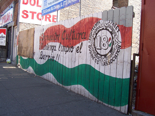 A wooden fence is painted in the colors of the Mexican flag, with the logo of the Eighteenth Street Business Association and the message 'Mantenga limpio el Barrio!!