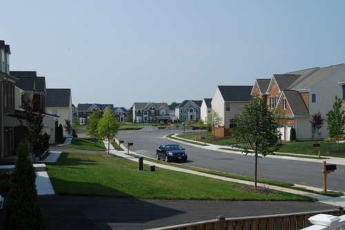 A gently curving residential street is flanked by rows of similar houses, each with a wide grassy front yard