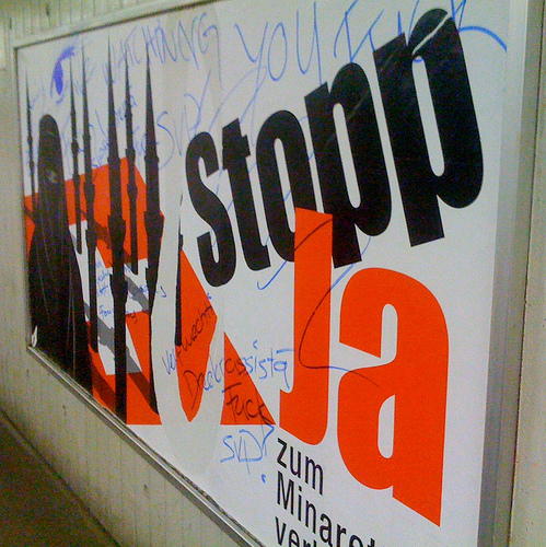 Poster showing black silhouettes of a woman in a burkha and numerous minarets on the Swiss flag, with the words 'Stopp Ja.' Blue pen graffiti has been scrawled over it.