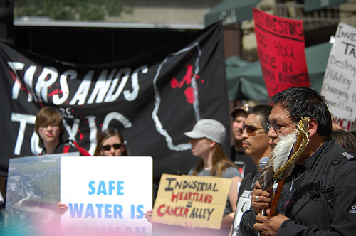 At the right of the image, a Native man holds a pipe with feathers on it and speaks into a microphone. A large black banner to his right reads 'tarsands are toxic.' Other protesters hold signs saying 'safe water is ...' and 'industrial heartland = cancer alley.'