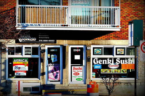 Two storefronts are seen in a brick building. The one on the left offers extensions and other hair styling, with a large poster advertising money transfers to Africa. The one on the right is Genki Sushi, with pictures of food in the window. Several leafless trees and construction barrels are in the foreground.