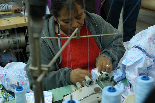 An older latina woman is seen at an angle from above as she sits at a sewing machine, working on a piece of blue flower-print fabric.